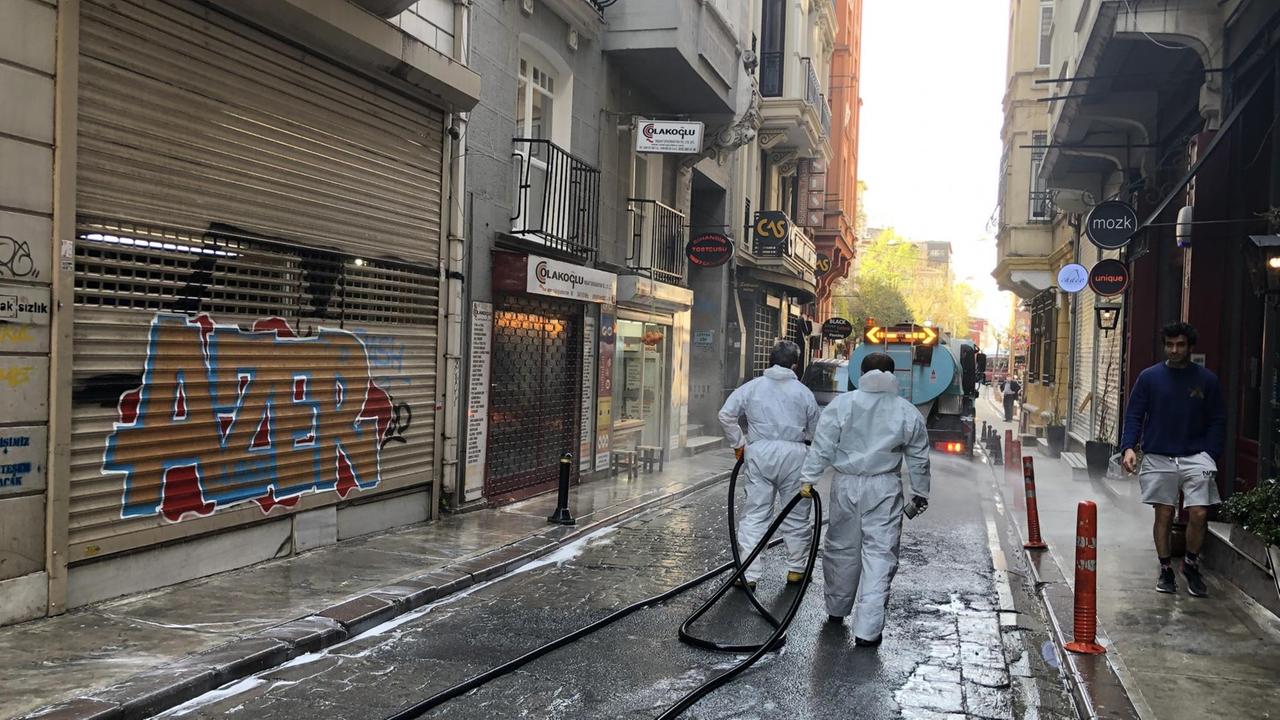 Zwei Männer mit weißer Schutzkleidung und langen Wasserschläuchen gehen durch eine nasse Gasse mit geschlossenen Läden in Istanbul. Sie folgen einem großen weißen Fahrzeug. Zwei Männer mit weißer Schutzkleidung und langen Wasserschläuchen gehen durch eine nasse Gasse mit geschlossenen Läden in Istanbul. Sie folgen einem großen weißen Fahrzeug.