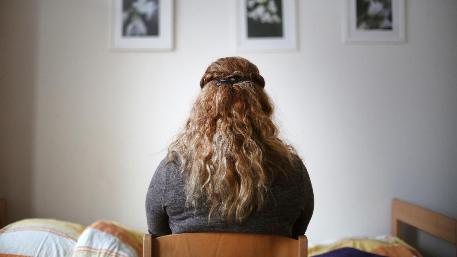 Eine Frau mit langen blonden gelockten Haaren sitzt mit dem Rücken zum Betrachter in einem Zimmer und schaut auf eine Wand mit drei Bildern Eine Frau mit langen blonden gelockten Haaren sitzt mit dem Rücken zum Betrachter in einem Zimmer und schaut auf eine Wand mit drei Bildern