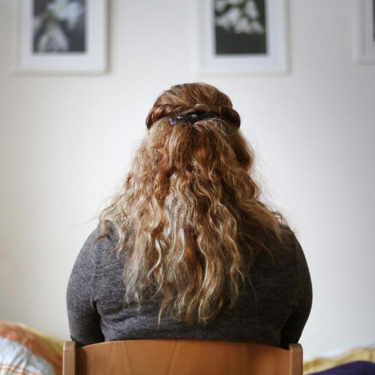 Eine Frau mit langen blonden gelockten Haaren sitzt mit dem Rücken zum Betrachter in einem Zimmer und schaut auf eine Wand mit drei Bildern