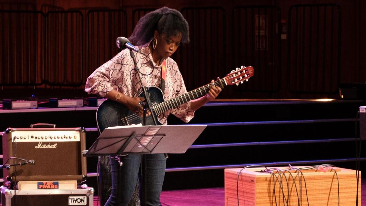Eine Frau in heller Bluse und schwarzen Locken hält eine Gitarre in der Hand. Sie steht zwischen einem Gitarrenverstärker und einem Notenständer. Eine Frau in heller Bluse und schwarzen Locken hält eine Gitarre in der Hand. Sie steht zwischen einem Gitarrenverstärker und einem Notenständer.