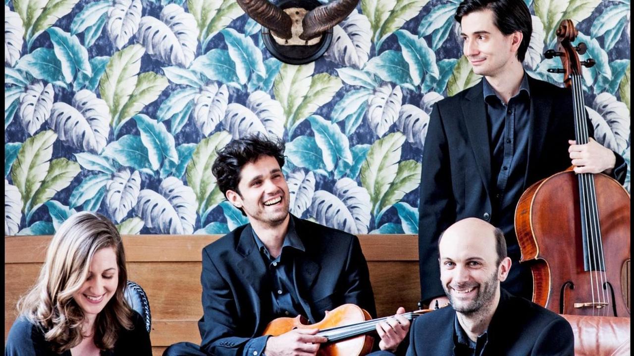 Die drei Männer und die die Frau des Quartetts sitzen mit ihren Instrumenten vor einer Wand, die mit einer floralen Tapete tapeziert ist und auf der ein Antilopengeweih prangt.