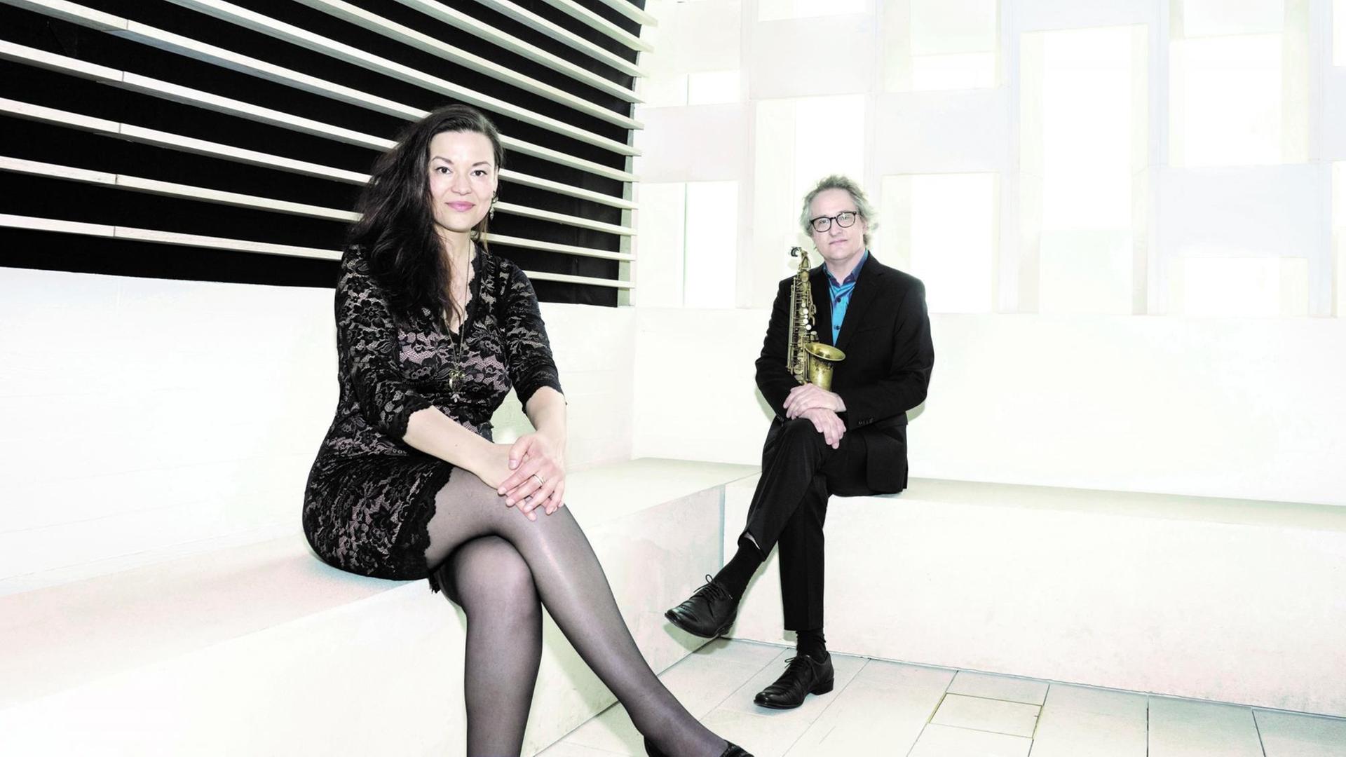 In der Ecke eines weißen Raumes sitzt im Vordergrund eine Frau im kurzen schwarzen Kleid, rechts im Hintergrund ein Mann im schwarzen Anzug und blauen Hemd. Beide lächeln. In der Ecke eines weißen Raumes sitzt im Vordergrund eine Frau im kurzen schwarzen Kleid, rechts im Hintergrund ein Mann im schwarzen Anzug und blauen Hemd. Beide lächeln.