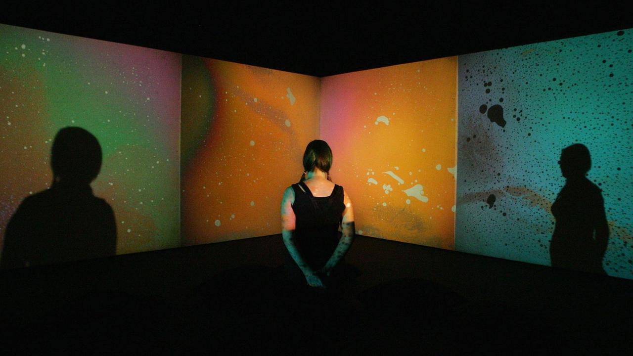 Eine Frau schaut das Werk 'Liquid Crystal Environment' von Gustav Metzger an. Im Hintergrund sieht man Farbflächen in Grün, Orange und Blau.