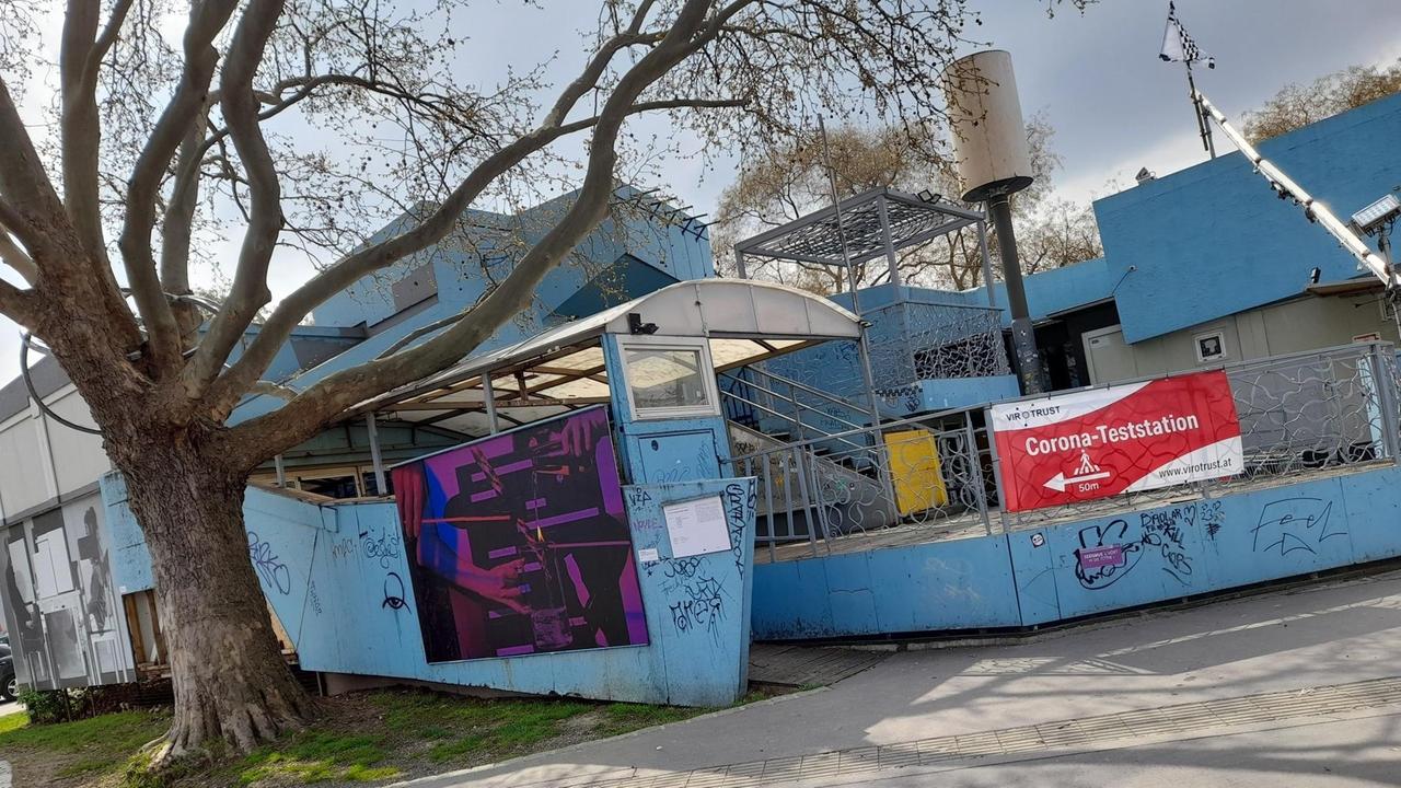 Das blaue Clubgebäude hat unssymetrische Formen. Am Eigang hängt ein rotes Schild mit der Aufschrift: "Corona-Teststation". Links neben dem Club steht ein riesiger Baum. Das blaue Clubgebäude hat unssymetrische Formen. Am Eigang hängt ein rotes Schild mit der Aufschrift: "Corona-Teststation". Links neben dem Club steht ein riesiger Baum.