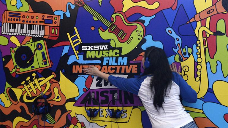 Eine Frau mit Kopfhörern auf den Ohren steht mit dem Rücken zum Betrachter vor einer bunt bemalten Wand auf der die Begriffe "SXSW", "Music", "Film", Interactive", "2018" und "Austin" zu lesen sind Eine Frau mit Kopfhörern auf den Ohren steht mit dem Rücken zum Betrachter vor einer bunt bemalten Wand auf der die Begriffe "SXSW", "Music", "Film", Interactive", "2018" und "Austin" zu lesen sind