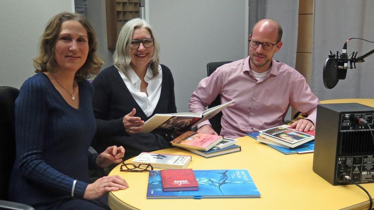 Tanya Lieske, Ute Wegmann und Jan Drees sitzen in einem Aufnahmestudio an einem Tisch und haben Bücher in der Hand. Tanya Lieske, Ute Wegmann und Jan Drees sitzen in einem Aufnahmestudio an einem Tisch und haben Bücher in der Hand.