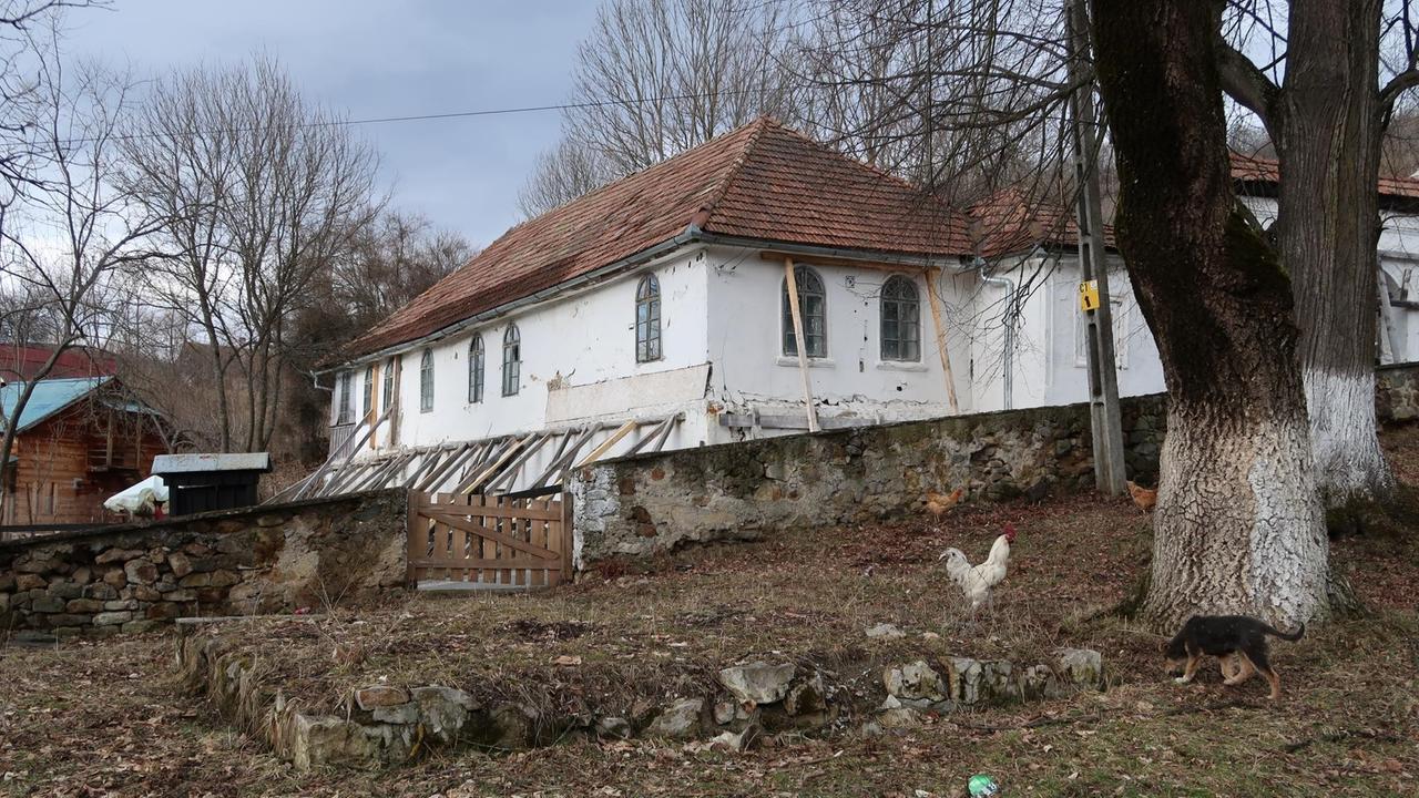 Seitliche Ansicht eines weißgestrichenen Gebäudes an einem Hang. Die Fensterscheiben sind zum Teil zersplittert und das Haus wird von Balken gestützt. Seitliche Ansicht eines weißgestrichenen Gebäudes an einem Hang. Die Fensterscheiben sind zum Teil zersplittert und das Haus wird von Balken gestützt.