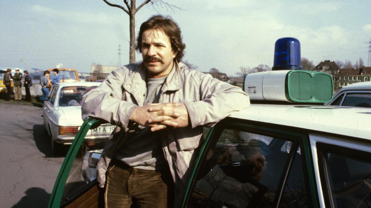Götz George steht bei den Dreharbeiten zum ersten Schimanski-Tatort im Jahr 1981 vor einem Polizeiauto.