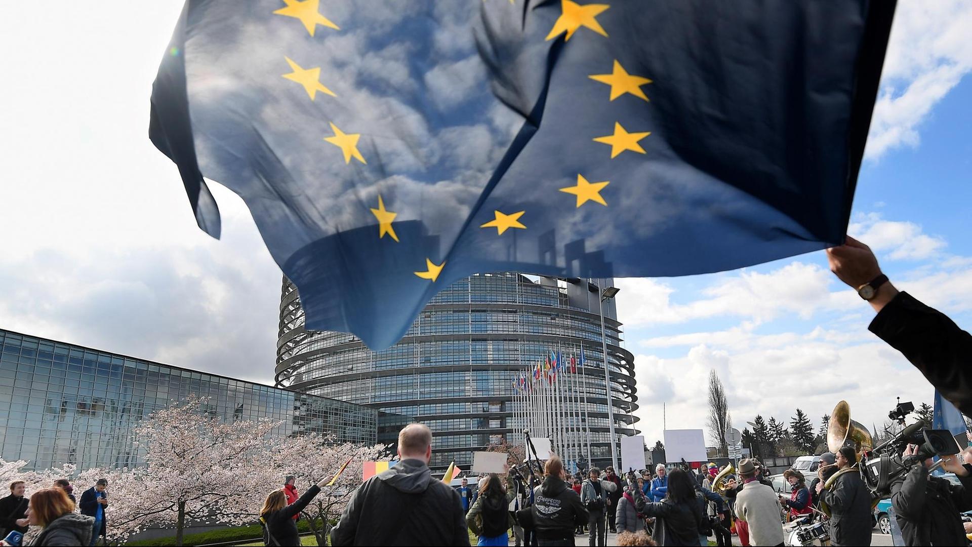 Menschen stehen vor dem runden Parlamentsgebäude in Straßburg. Vorne im Bild schwenkt jemand eine EU-Flagge. Menschen stehen vor dem runden Parlamentsgebäude in Straßburg. Vorne im Bild schwenkt jemand eine EU-Flagge.