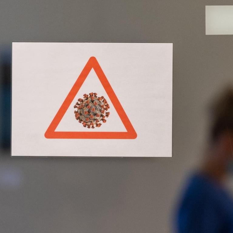 Eine Gesundheits- und Krankenpflegerin läuft auf der Corona-Station im Städtischen Klinikum Dresden in Schutzausrüstung hinter einer Scheibe an der ein Achtungssymbol mit einem stilisierten Cornavirus angebracht ist vorbei. 