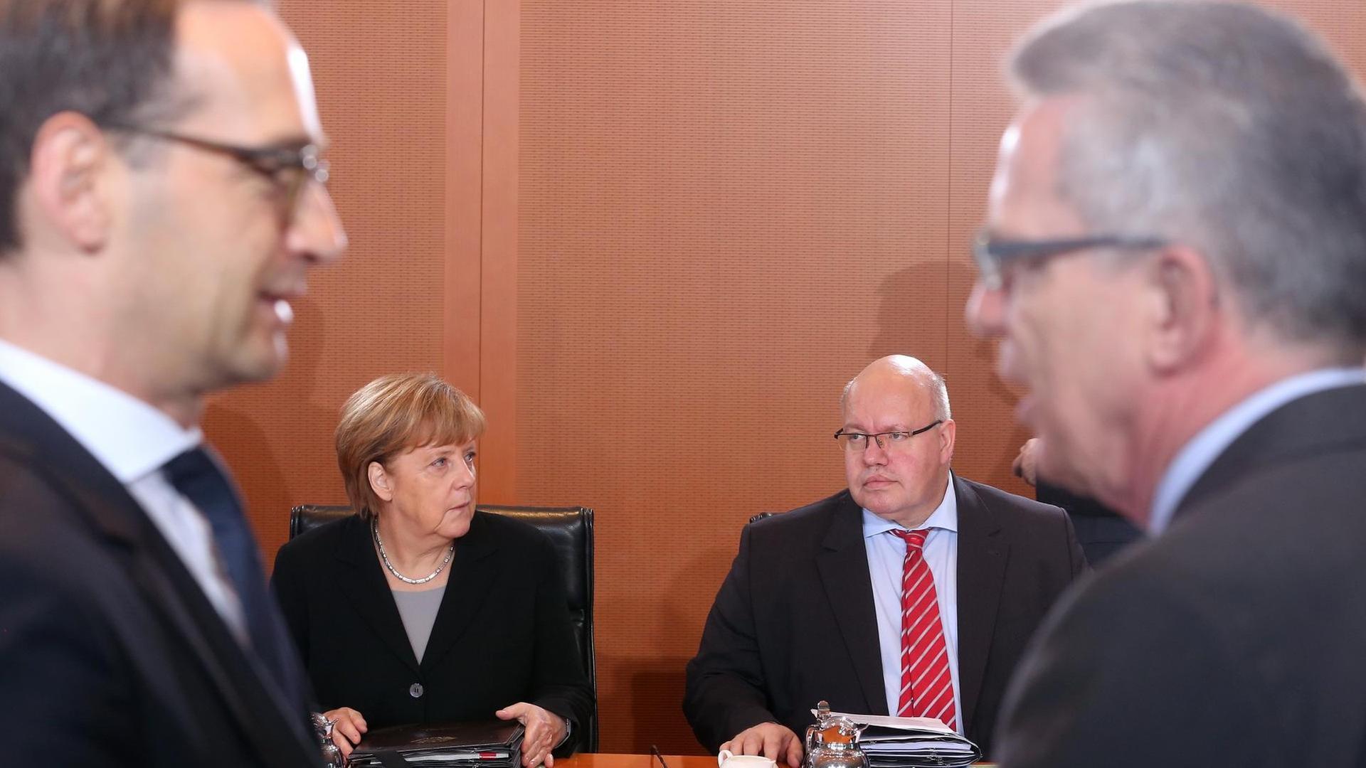 Das Bild zeigt Maas und de Maiziere im Vordergrund und unscharf stehend am linken und rechten Bildrand, die miteinander reden. In der Bildmitte sind im Hintergrund und scharf Merkel und Altmaier sitzend am Kabinettsstisch zu sehen.