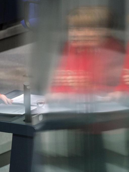 Bundeskanzlerin Angela Merkel (CDU) steht in einem roten Blazer am Rednerpult des Bundestags. Ihr Bild spiegelt sich in einer Scheibe.