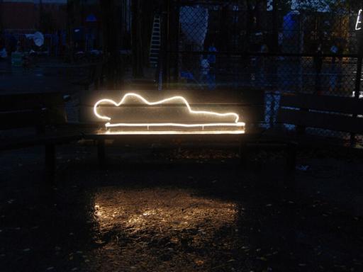 Vor einem dunklen Hintergrund ist eine Neonskulptur zu sehen: die leuchtende Silhouette einer obdachlosen Person, die auf einer Matratze liegt