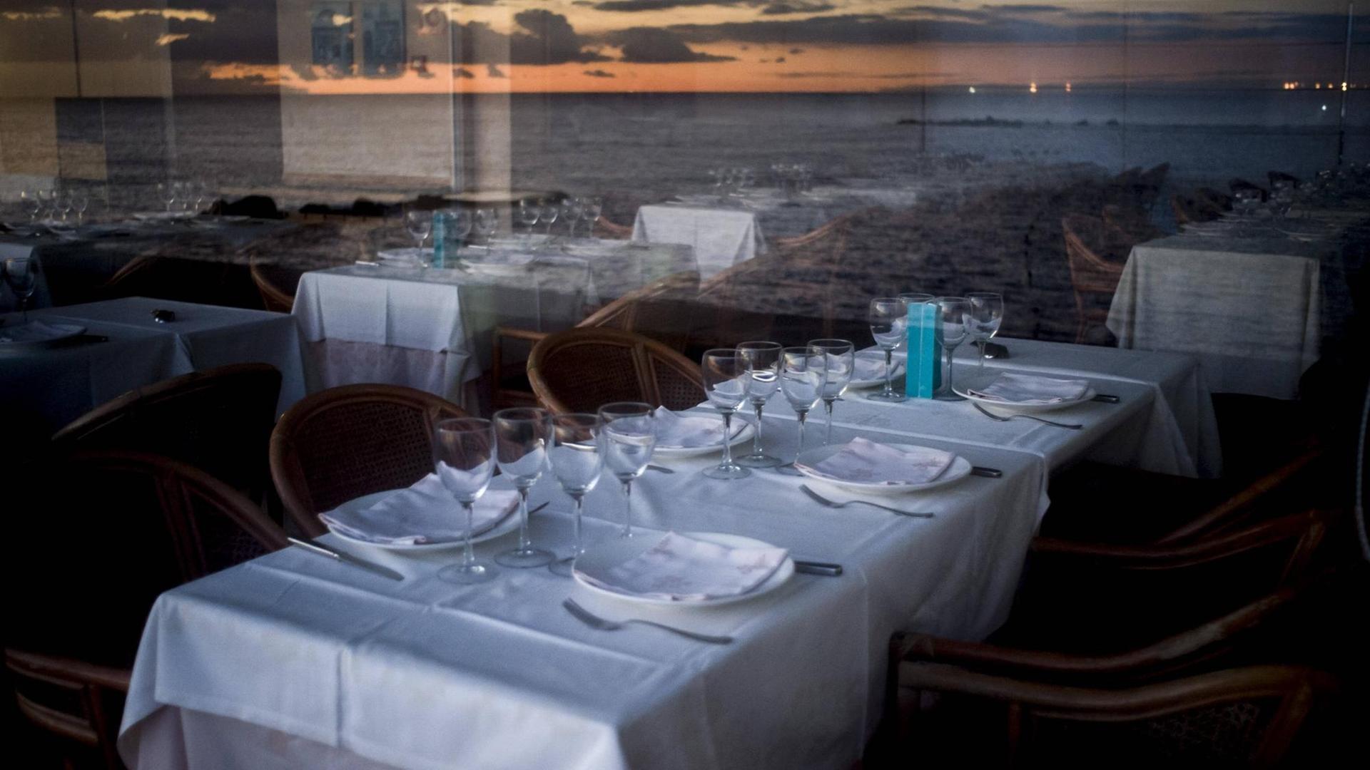 Leere Tische in einem Restaurant, in den Fensterscheiben spiegelt sich die Küste.
