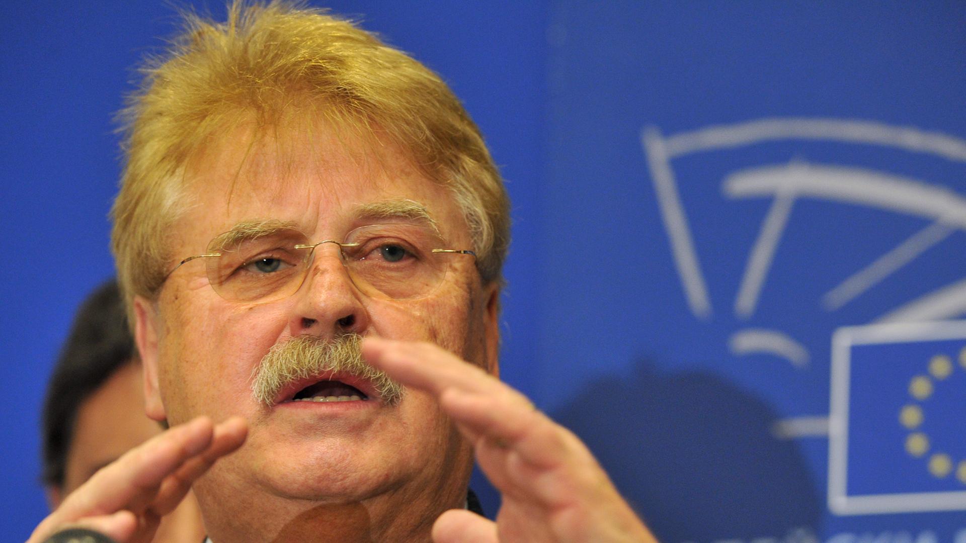 Elmar Brok spricht vor einem Mikrofon, mit den Händen gestikulierend, vor blauem Hintergrund mit weißer kyrillscher Aufschrift. Elmar Brok spricht vor einem Mikrofon, mit den Händen gestikulierend, vor blauem Hintergrund mit weißer kyrillscher Aufschrift.