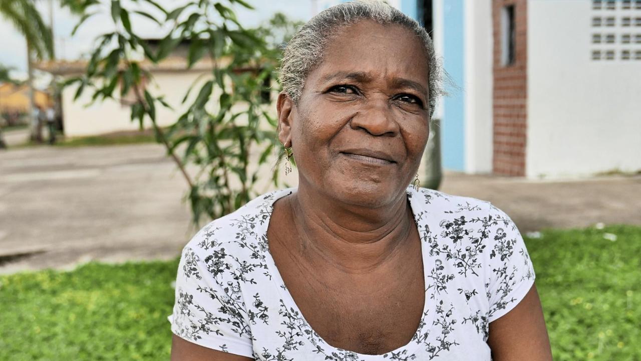 Macária Allin, eine Afrokolumbianerin mit grau-schwarzem Stoppelhaar und weißem Top, sitzt draußen im Grünen und schaut freundlich in die Kamera. Macária Allin, eine Afrokolumbianerin mit grau-schwarzem Stoppelhaar und weißem Top, sitzt draußen im Grünen und schaut freundlich in die Kamera.