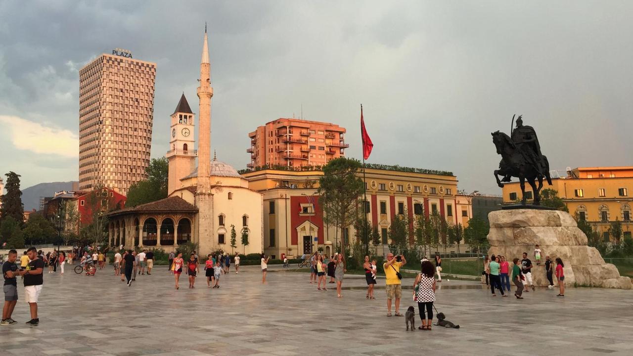 Menschen laufen über den Skanderbergplatz in Tirana. Keiner trägt Kopftuch. Ein Minarett einer Moschee ist zu sehen.