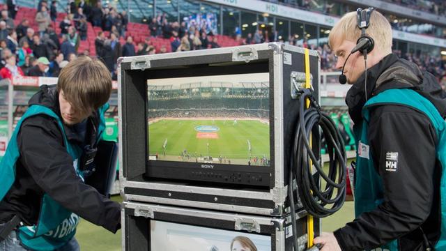 Zwei Männer stehen links und rechts von einem Fernseher, auf dem ein Fußball-Spiel übertragen wird.