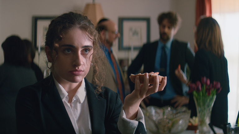 Im Still aus Emma Seligmans "Shiva Baby" hält die Protagonistin auf einer Trauerfeier ein belegtes Brötchen in der Hand. Im Still aus Emma Seligmans "Shiva Baby" hält die Protagonistin auf einer Trauerfeier ein belegtes Brötchen in der Hand.