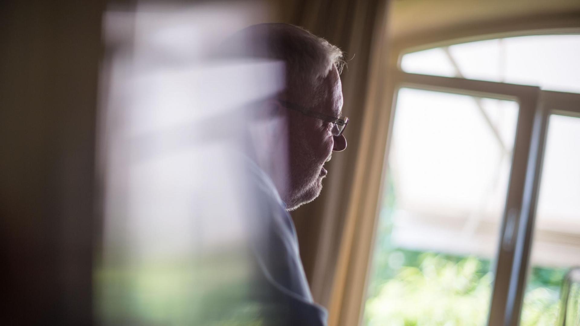 Auf dem Foto ist ein älterer Mann im Profil zu sehen, der aus einem Fenster schaut. Auf dem Foto ist ein älterer Mann im Profil zu sehen, der aus einem Fenster schaut.