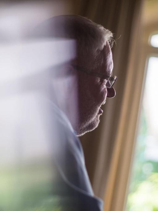Auf dem Foto ist ein älterer Mann im Profil zu sehen, der aus einem Fenster schaut.