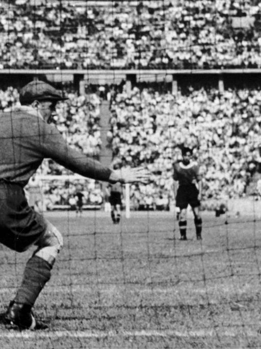 Blick auf ein Tor von hinten. Man sieht wie der Ball im Tor landet und der Torwart sich noch in die Richtung des Balles streckt. Der Torwart trägt eine Schiebermütze. (Historische Fotografie)