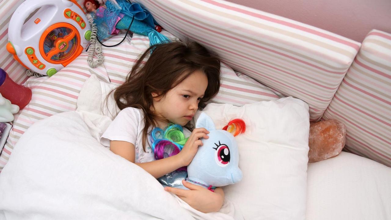Ein kleines Mädchen liegt in einem Kinderzimmer und kuschelt mit ihrem Plüschtier (gestellte Szene).