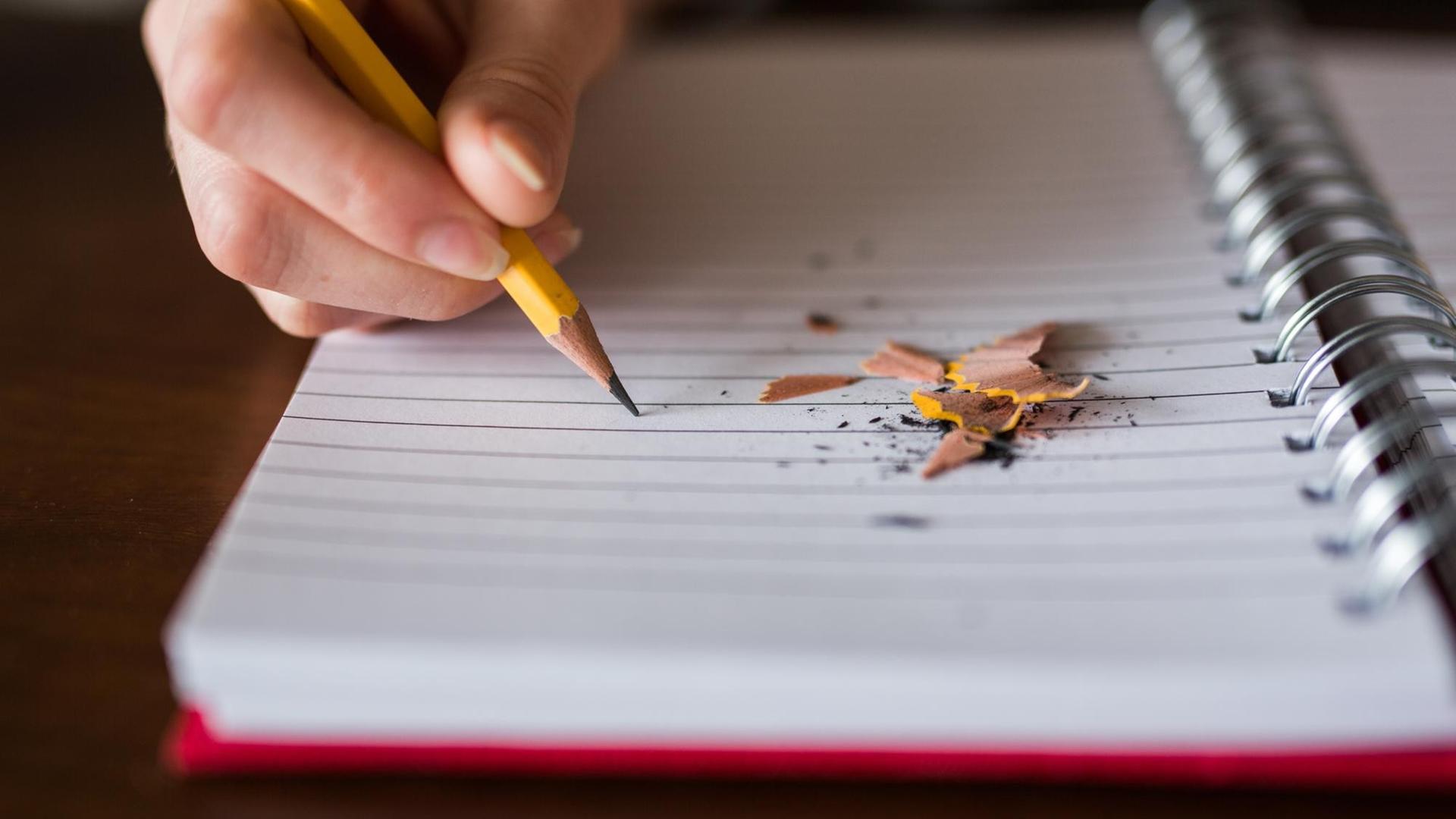 Eine Hand schreibt mit gelbem Bleistift auf einem Block, daneben liegen die Reste des angespitzen Bleistifts. Eine Hand schreibt mit gelbem Bleistift auf einem Block, daneben liegen die Reste des angespitzen Bleistifts.