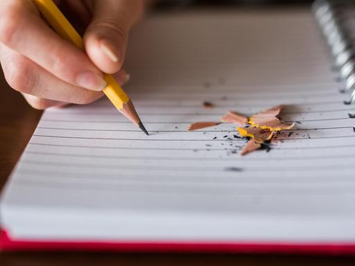 Eine Hand schreibt mit gelbem Bleistift auf einem Block, daneben liegen die Reste des angespitzen Bleistifts.