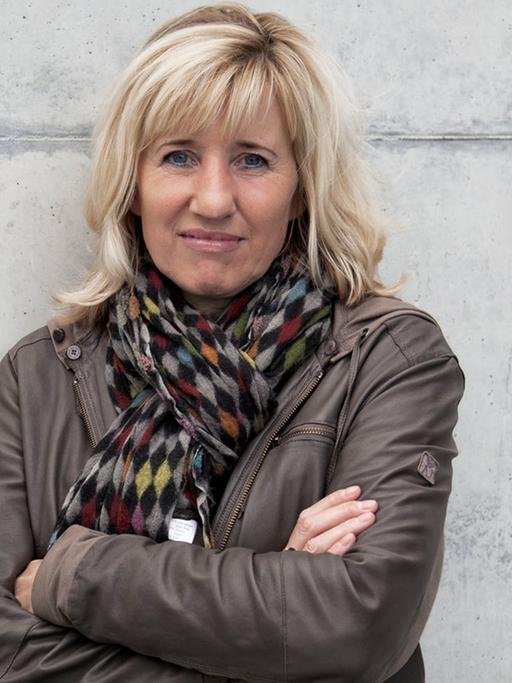 Die Schriftstellerin und Doping-Expertin Ines Geipel, die Arme verschränkt, an eine graue Betonmauer gelehnt.