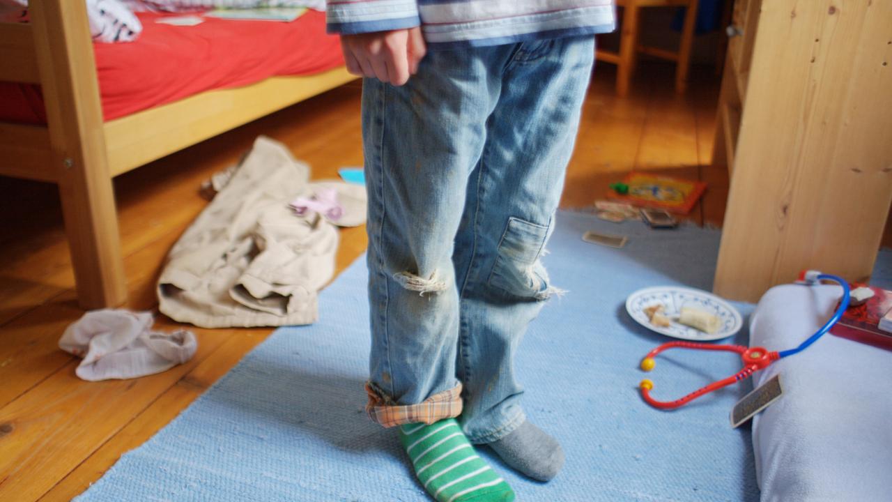 Ein kleiner Junge steht mit schmutziger Kleidung, zerrissenen Hosen und verschiedenen Strümpfen in einem unaufgräumten Zimmer.
