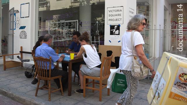 Riock Lowe sitzt mit drei Menschen um einen kleinen Holztisch am Rande einer Straße. Im Hintergrund ist ein verglastes Ladenblokal zu erkennen, das die Werkstatt für sein documenta 14-Projekt "Victoria Square Project" bildet.