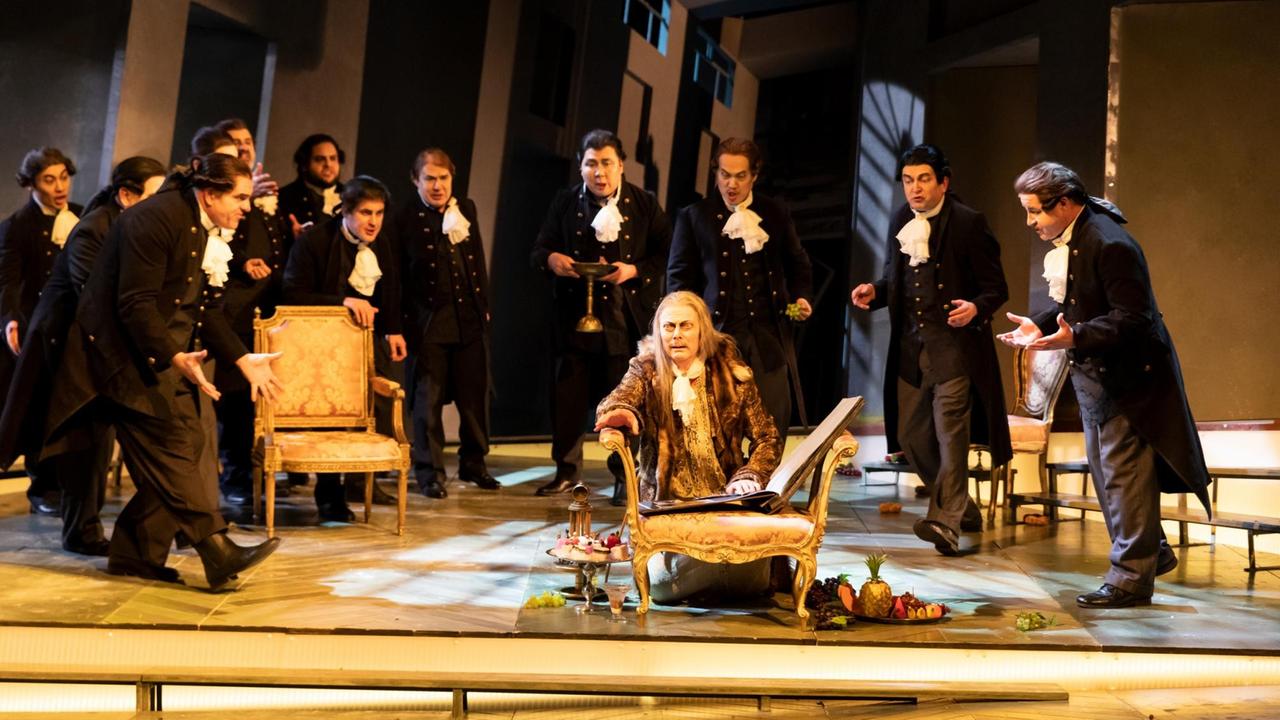 Ein Mann kniet auf dem Boden und blättert in einem Album. Um ihn herum sind schwarz gekleidete Männer mit weißen Halstüchern. Szene aus "Das Schloss Dürande" am Staatstheater Meiningen. Ein Mann kniet auf dem Boden und blättert in einem Album. Um ihn herum sind schwarz gekleidete Männer mit weißen Halstüchern. Szene aus "Das Schloss Dürande" am Staatstheater Meiningen.