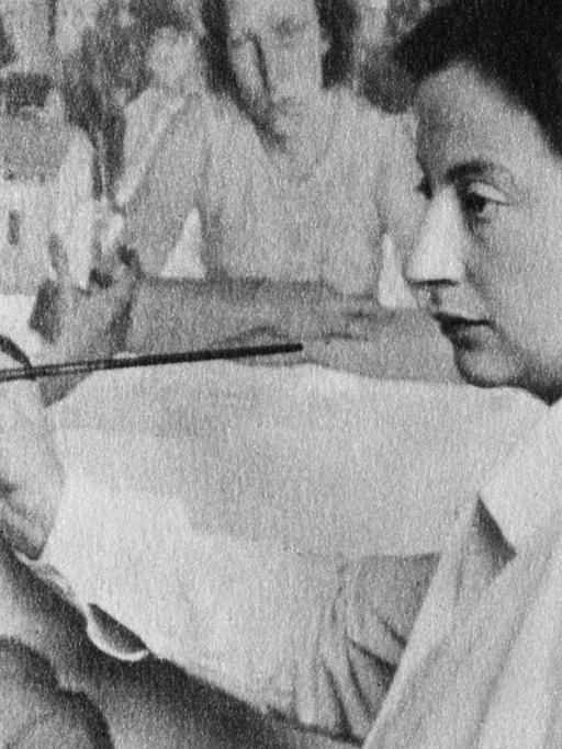 Schwarzweiß Fotografie: Lotte Laserstein steht vor einem Gemälde mit Pinsel und Farben. Sie trägt einen weißen Kittel.