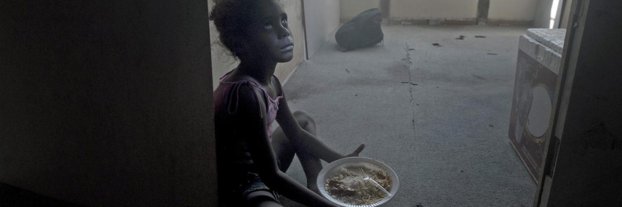Ein Kind sitzt auf dem Boden und hält einen Teller mit Essen in der Hand. 