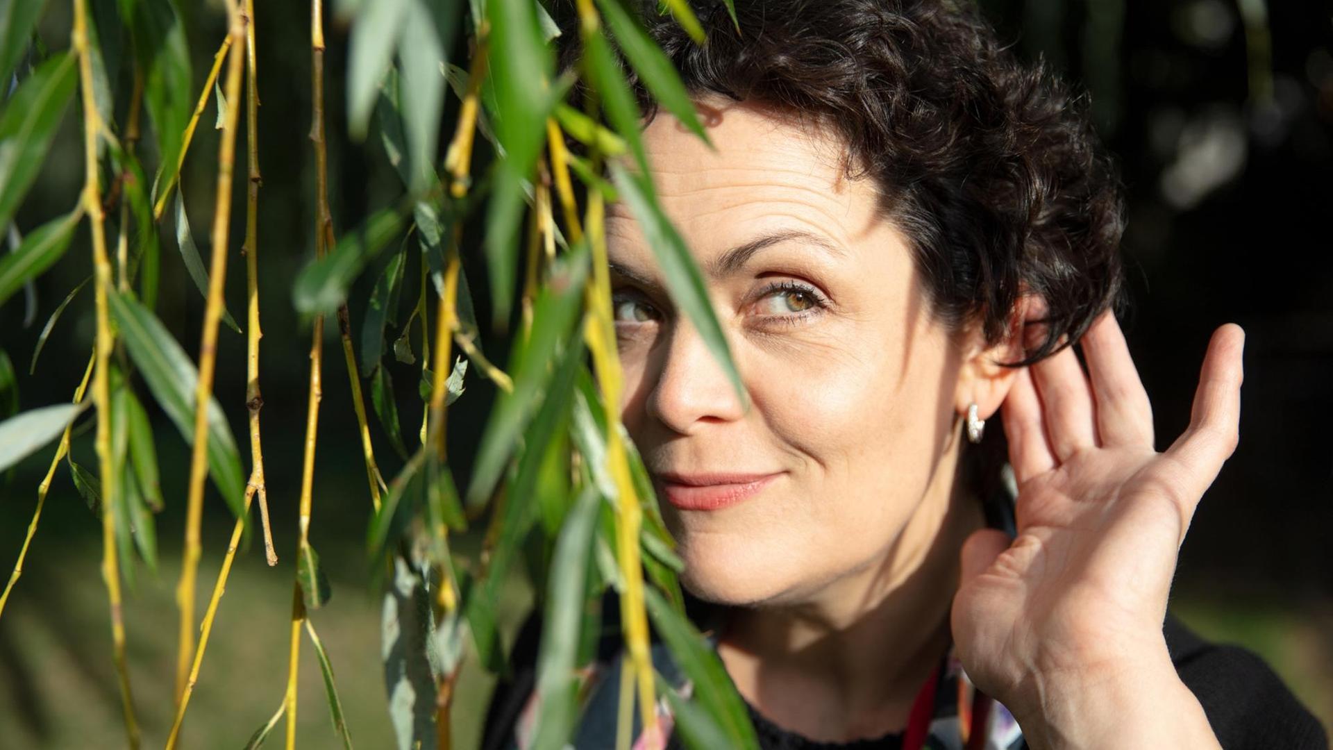 Ein Porträt der Bratschistin Tabea Zimmermann, Berlin 2019: Die Musikerin steht hinter herunterhängenden Ästen und horcht mit einer Hand am Ohr.