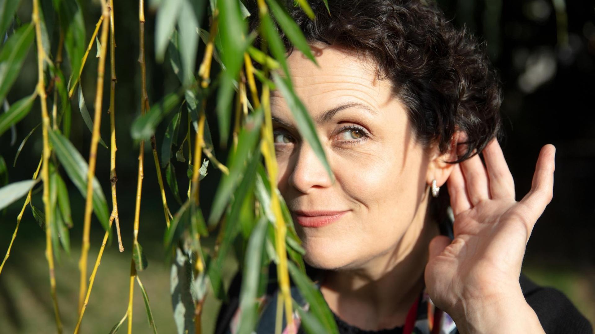 Ein Porträt der Bratschistin Tabea Zimmermann, Berlin 2019: Die Musikerin steht hinter herunterhängenden Ästen und horcht mit einer Hand am Ohr. Ein Porträt der Bratschistin Tabea Zimmermann, Berlin 2019: Die Musikerin steht hinter herunterhängenden Ästen und horcht mit einer Hand am Ohr.