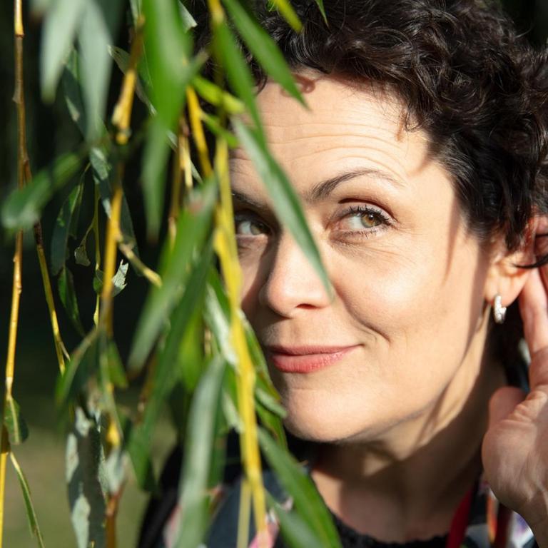 Ein Porträt der Bratschistin Tabea Zimmermann, Berlin 2019: Die Musikerin steht hinter herunterhängenden Ästen und horcht mit einer Hand am Ohr.
