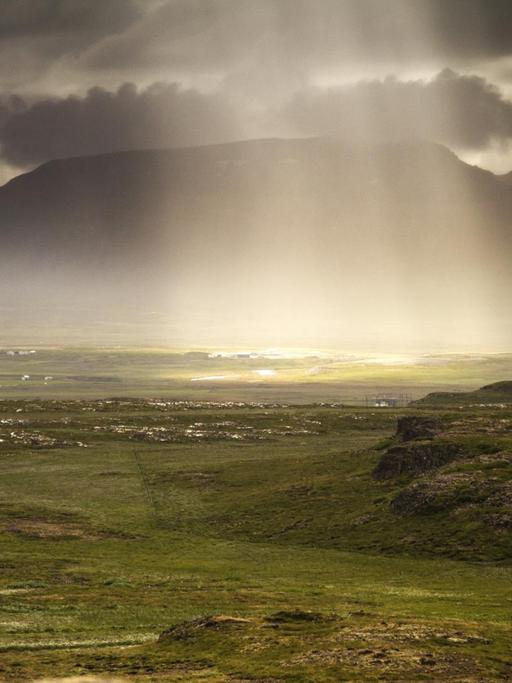 Sonnenlicht fällt durch die dichte, graue Wolkendecke auf die Landschaft Islands.