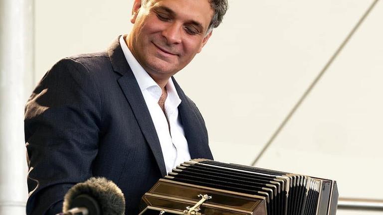 Ein Mann sitzt mit einem Bandoneon auf den Knien und spielt, während er auf das Mikrophon neben ihm schaut, das seine Musik aufzeichnet. Ein Mann sitzt mit einem Bandoneon auf den Knien und spielt, während er auf das Mikrophon neben ihm schaut, das seine Musik aufzeichnet.