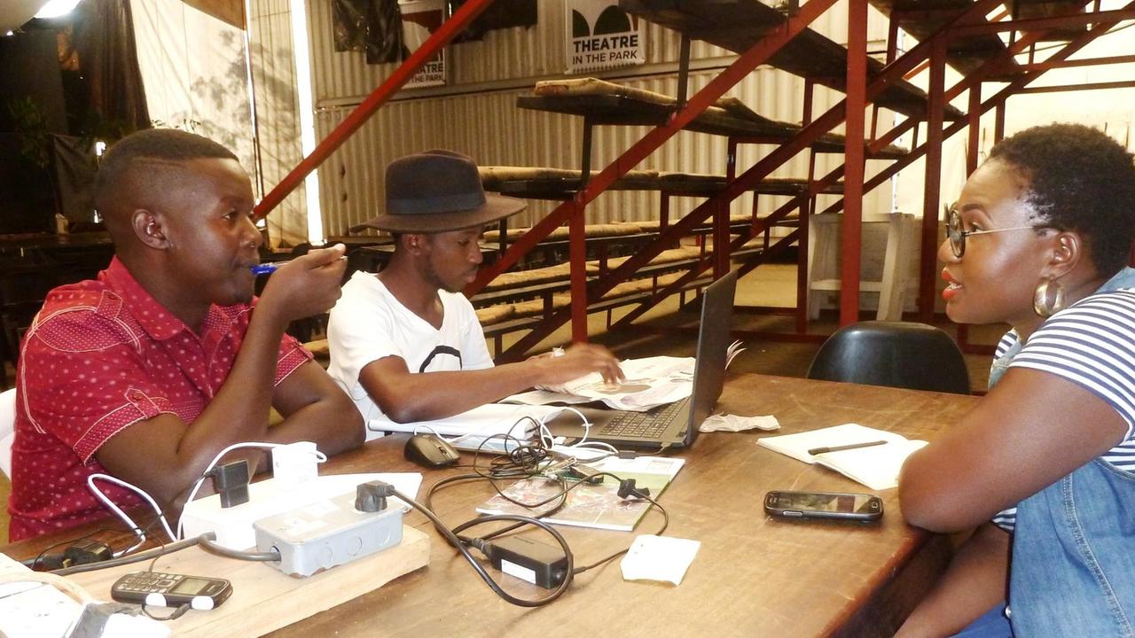 Victor Mpofu alias Doc Vikela (l.) und sein Team sitzen am Tisch mit Laptops und Handys und sind beim Brainstorming für ihre Comedy-TV-Show