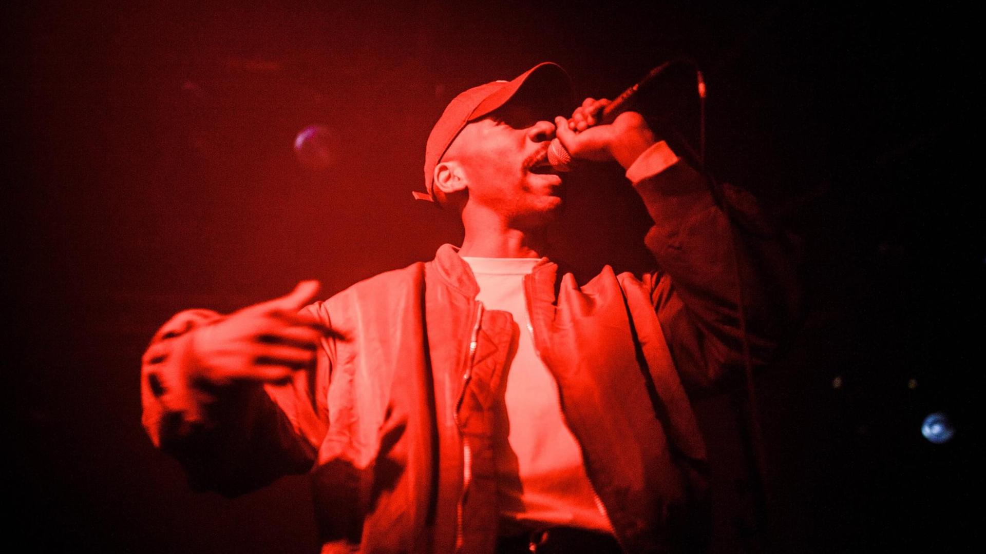 Musiker Dean Blunt in rotes Licht getaucht, mit Mikrofon auf der Bühne. Musiker Dean Blunt in rotes Licht getaucht, mit Mikrofon auf der Bühne.