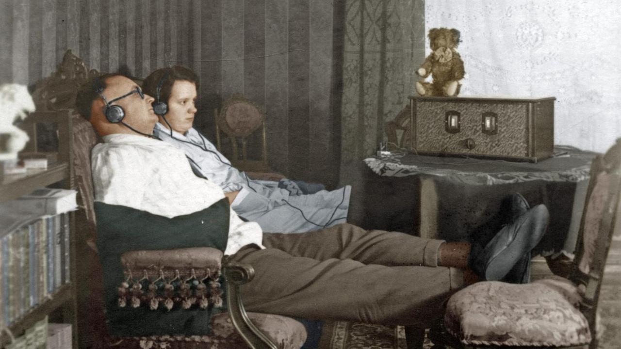 Ein Mann und eine Frau sitzen nebeneinander auf dem Sofa und hören mit Kopfhörern Radio, digital kolorierte Aufnahme aus dem Jahr 1928 Ein Mann und eine Frau sitzen nebeneinander auf dem Sofa und hören mit Kopfhörern Radio, digital kolorierte Aufnahme aus dem Jahr 1928