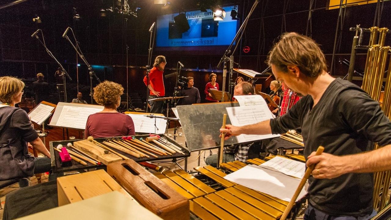 Mehrere Musikerinnen und Musiker sind von hinten mit ihren Instrumenten abgelichtet. Sie sitzen und stehen auf der einer Bühne. Im Hintergrund ist eine Leinwand auf der zu lesen ist: Deutschlandfunk Kammermusiksaal. Mehrere Musikerinnen und Musiker sind von hinten mit ihren Instrumenten abgelichtet. Sie sitzen und stehen auf der einer Bühne. Im Hintergrund ist eine Leinwand auf der zu lesen ist: Deutschlandfunk Kammermusiksaal.