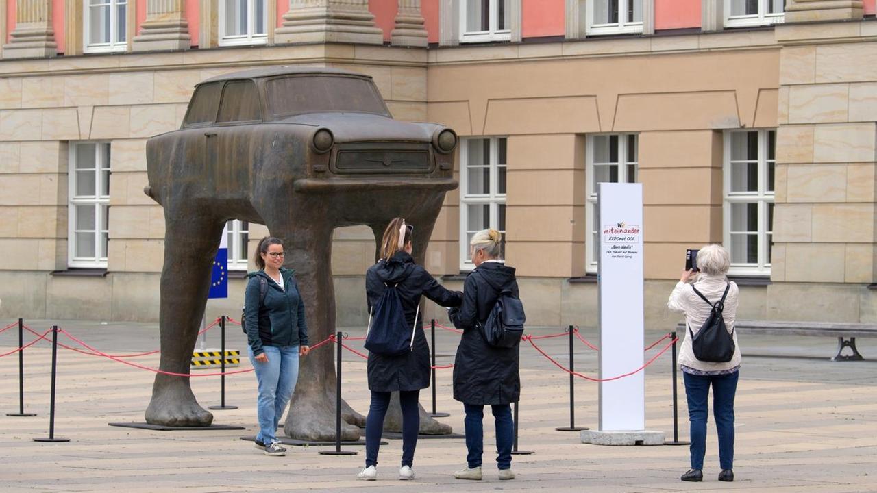 Passanten stehen vor der Skulptur eines Trabant auf Beinen.