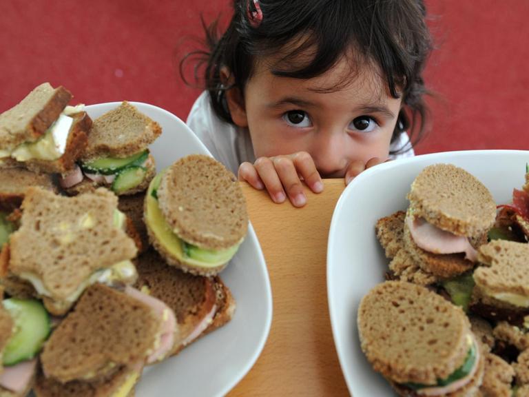 Ein kleines Kind schaut in die Kamera. Im Vordergrund stehen zwei Teller mit Pausenbroten, ausgestochen in unterschiedlichen Formen und belegt mit Gemüse, Käse und Aufschnitt.