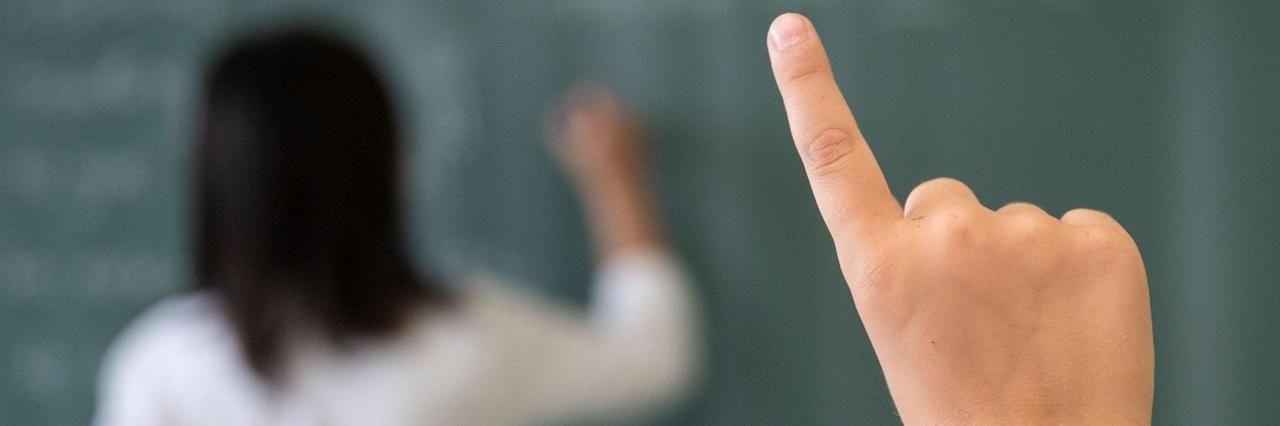 Hand eines Schülers einer fünften Klasse, der sich während einer Unterrichtsstunde meldet. Im Hintergrund des Bildes schreibt die Lehrerin an die Tafel. 