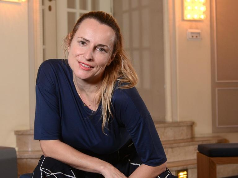 Anna Bergmann, Schauspieldirektorin des Badischen Staatstheaters. Sie sitzt im Theaterfoyer auf einem Hocker und schaut direkt in die Kamera. Foto: Mike Wolff | Verwendung weltweit