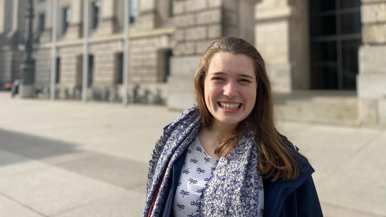 Eine junge Frau blickt breit lächelnd in die Kamera. Sie trägt ein blau-weiß gemustertes T-Shirt, einen Schal und eine blaue Jacke und steht vor einem historischen Gebäude. Es handelt sich um die Politikerin Emilia Fester. Eine junge Frau blickt breit lächelnd in die Kamera. Sie trägt ein blau-weiß gemustertes T-Shirt, einen Schal und eine blaue Jacke und steht vor einem historischen Gebäude. Es handelt sich um die Politikerin Emilia Fester.