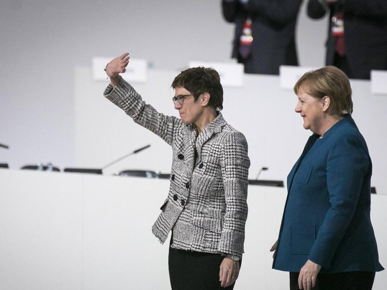Annegret Kramp-Karrenbauer winkt ins Publikum, rechts neben ihr steht Angela Merkel in lockerer Pose